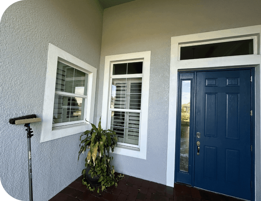 Front porch with a blue door, two windows, a potted plant, and a cleaning tool leaning against the wall.