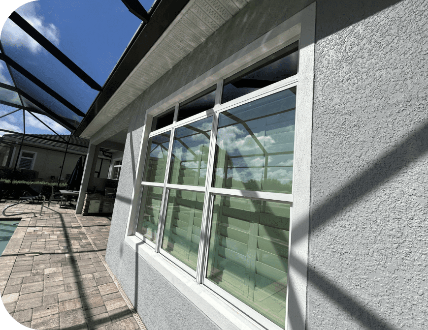 Sunlit house exterior with large windows reflecting clouds, beside a patio and pool area. Shadows from an overhead structure create lines on the walls and paving stones.