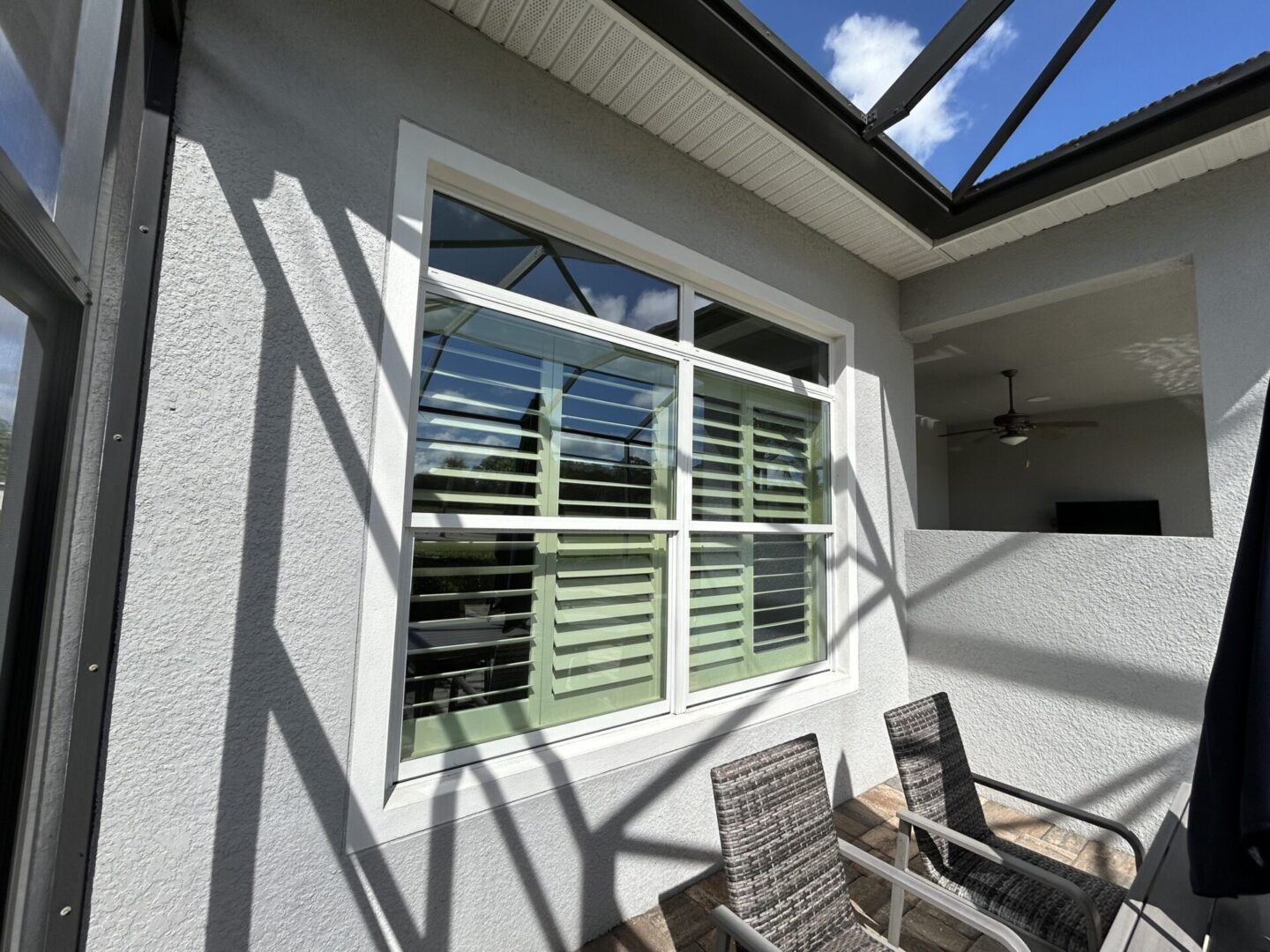 Outdoor patio area with white-framed window and two wicker chairs. Sunlight casts geometric shadows on the wall. Ceiling fan visible inside through an open doorway.