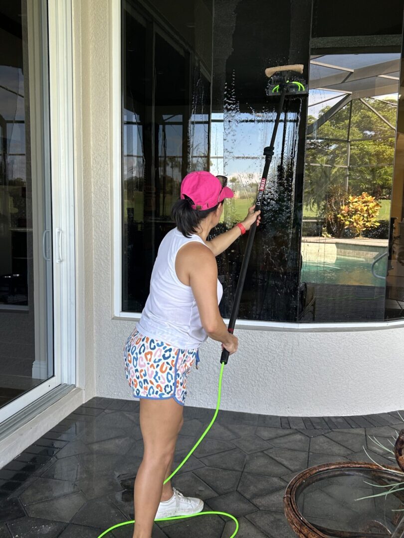 A person in a pink cap and patterned shorts uses a long-handled brush to clean tall windows on a patio.