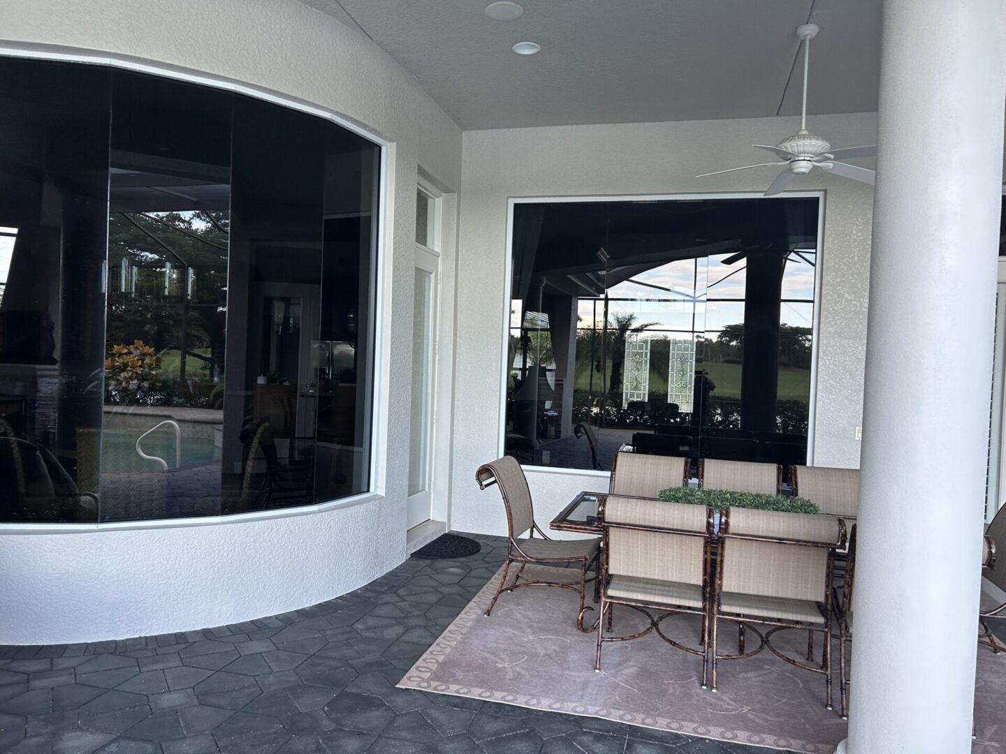 Patio with a dining table and chairs, protected by a covered area. Large windows reflect an outdoor view.