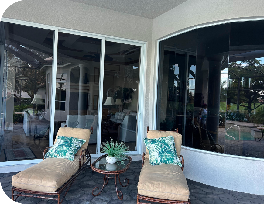 Two patio loungers with cushions and leaf-patterned pillows face a glass window, next to a small round table with a potted plant.