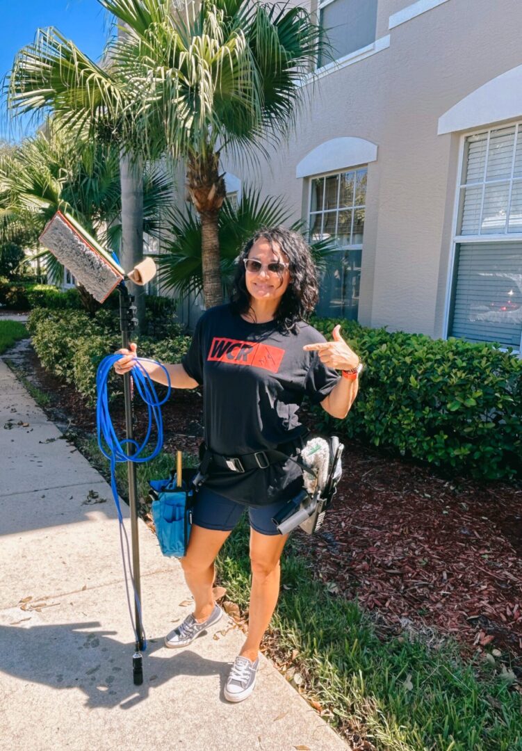 Woman stands outside, holding a cleaning tool and hose. She wears a black shirt, shorts, sunglasses, and sneakers. A residential building with greenery is in the background.